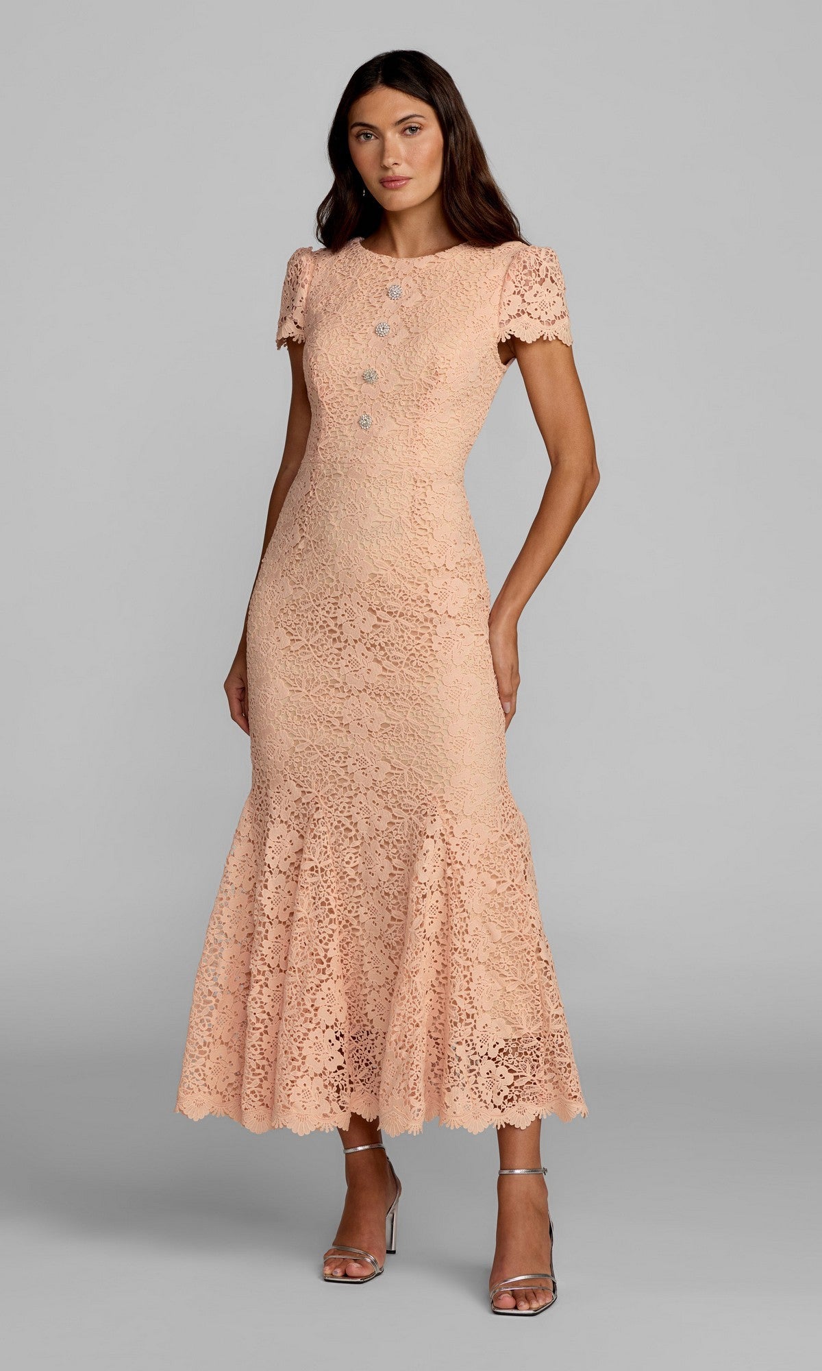 A woman stands against a plain gray background wearing the Mac Duggal 50116 tea-length party dress in peach, featuring short sleeves, silver buttons, an ankle-length skirt, and a ruffled hem—embodying refined elegance—paired with silver heeled sandals.